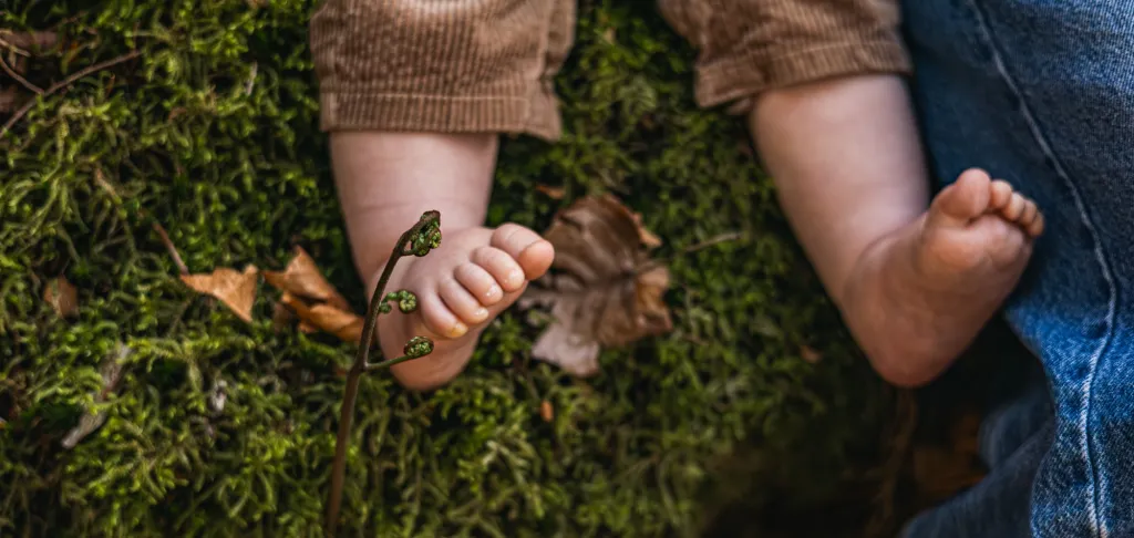 Olivia Greenland sits grounded against a mossy tree in a sacred forest grove, her posture open, expression calm—embodying the essence of ease, unmasking, and earth-rooted witnessing.